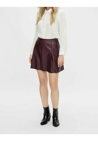 Jupe en cuir bordeaux avec plis, associée à une chemise blanche à boutons. Le modèle porte des bottines noires à la cheville. Design épuré et minimaliste.