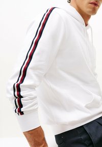 Sudadera blanca con un bolsillo frontal, que presenta rayas rojas y marinas en las mangas y puños de canalé. Textura de tela suave, ajuste casual.