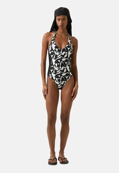 Maillot de bain une pièce noir et blanc, mettant en avant un motif géométrique, un design à encolure halter et des panneaux latéraux. Comprend un bonnet assorti et des tongs.