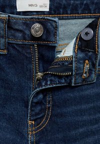 Jeans de mezclilla de un azul oscuro con un botón plateado y cremallera. Costuras naranjas visibles y detalle de forro negro en la cintura. Etiqueta de tamaño incluida.