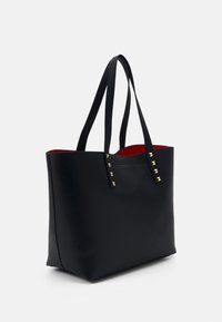 Sac fourre-tout noir en matériau lisse, doté d'un intérieur rouge, de deux longues poignées et de clous décoratifs dorés sur un côté.