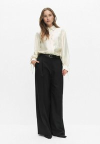 Crème satijnen blouse met knoopsluiting en manchetdetails, gecombineerd met hoge zwarte wijde broek en een zwarte riem. Zwarte puntige schoenen.