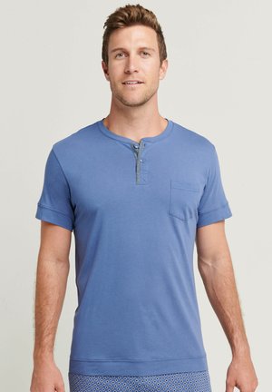 Mann mit heller Haut und kurzem braunem Haar, der ein blaues Henley-Shirt mit kurzen Ärmeln und Brusttasche trägt, steht vor einem schlichten Hintergrund.