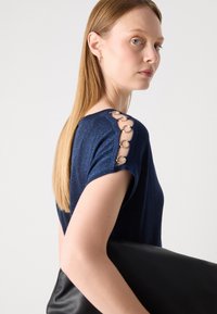Navyblaues kurzärmeliges Oberteil mit einem glitzernden Muster und metallischen Ringdetails an der Schulter. Das Model hält eine schwarze Clutch.