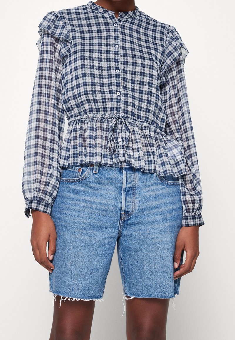 Personne portant un chemisier à carreaux bleu et blanc boutonné avec manches longues et un short en jean aux bords effilochés.