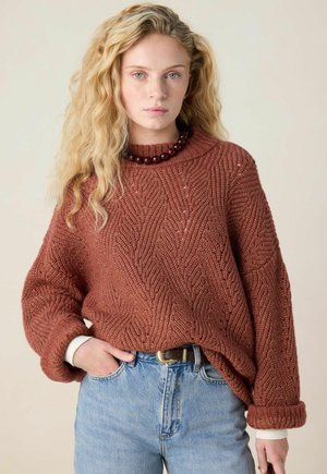 Young woman with long wavy blonde hair wearing a rust-colored knit sweater, blue jeans, a belt, and dark beaded necklace stands against plain background.