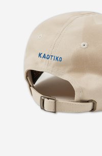 Gorra beige con visera curvada, correa ajustable y bordado azul "KAOTIKO" en la parte posterior. La textura de la tela parece resistente y suave.
