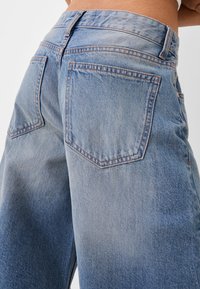 Lähikuva henkilöstä, joka pukeutuu vaaleansinisiin korkeavyötäröisiin farkkuihin, joissa näkyvät takataskut ja vyötärö, yksinkertaista taustaa vasten.