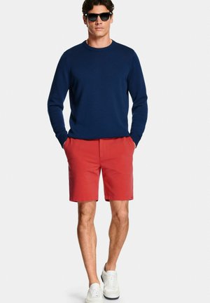 Man met zwarte zonnebril, marineblauwe trui met lange mouwen, rode korte broek en witte sneakers tegen een effen achtergrond.