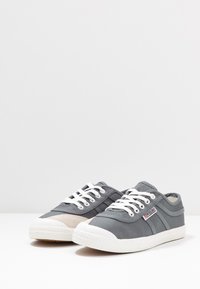 Sneaker di tela grigie con suole in gomma bianche, caratterizzate da un design con lacci, una striscia decorativa e una punta strutturata.