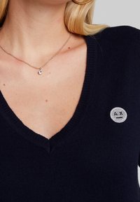 Navy v-neck sweater in soft knit fabric featuring a circular silver logo patch on the chest. Delicate chain necklace with a small pendant.