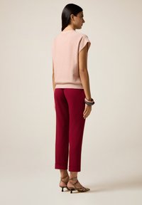 Vrouw die zijwaarts staat, gekleed in een lichtroze mouwloos shirt, burgundy broek tot de enkels, patroonhakken en drie armbanden om haar pols.