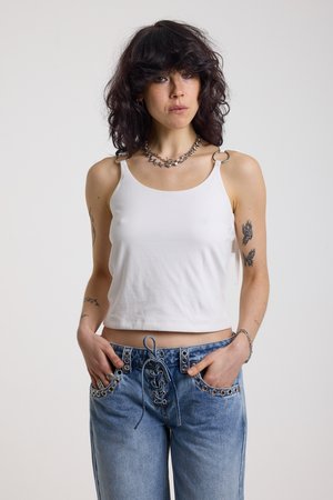 Γυναίκα με σγουρά σκούρα μαλλιά, ντυμένη με λευκό αμάνικο τοπ με λεπτομέρειες κρίκων και μπλε τζιν με κορδόνια στο μπροστινό μέρος και μεταλλικά στόμια, με τα χέρια στις τσέπες.