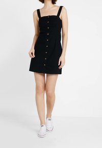 Frau in einem ärmellosen schwarzen Mini-Kleid mit Knopfleiste und breiten Trägern, die weiße Sneakers trägt, vor einem schlichten weißen Hintergrund stehend.