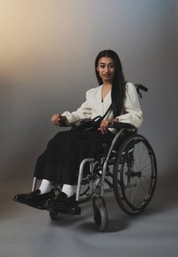 Una persona in sedia a rotelle con una blusa bianca e una gonna nera con texture a pieghe, che indossa scarpe nere e calzini bianchi, seduta su uno sfondo neutro.