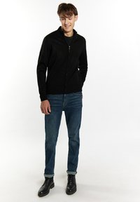 Svart zip-up tröja med ståkrage, texturerat stickat tyg, i kombination med blå jeans och svarta stövlar. Modellen står med händerna i fickorna.