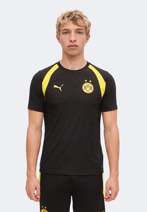 Jeune homme portant un maillot de football noir et jaune du Borussia Dortmund et un short assorti, debout devant un fond uni.