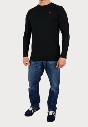 Chemise noire à manches longues avec un petit logo rouge sur la poitrine, associée à un jean bleu et des baskets noires avec des lacets blancs.