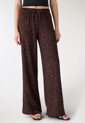 Donna che indossa pantaloni larghi a righe zebrate marroni e nere con top corto nero e sandali neri su sfondo bianco.