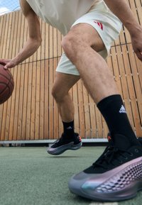 Sorte og grå treningssko med en teksturert overflate, en reflekterende detalj, og en gjennomsiktig såle. Spiller som dribler en basketball på banen.
