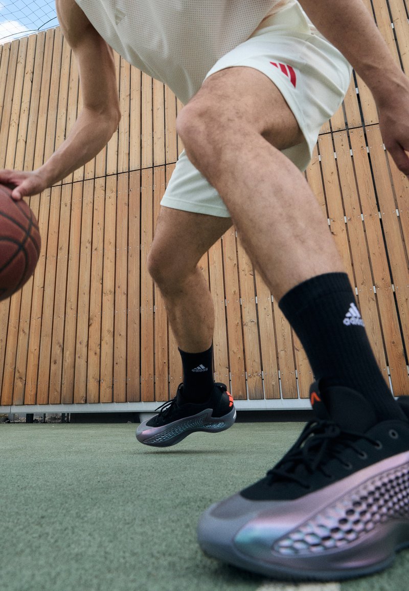 Sorte og grå treningssko med en teksturert overflate, en reflekterende detalj, og en gjennomsiktig såle. Spiller som dribler en basketball på banen.