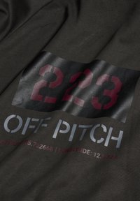 Czarna tkanina z dużymi, wyraźnymi cyframi "223" w kolorze bordowym, z napisem "OFF PITCH" i współrzędnymi w kolorze szarym. Teksturowana powierzchnia z delikatnym połyskiem.