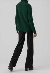 Persona vestida con un suéter de punto de tortuga verde oscuro, pantalones de pierna amplia negros y zapatos de tacón negros, de pie frente a un fondo gris liso.