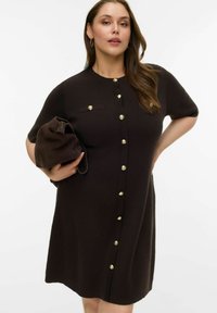 Põlveni ulatuv pruun kootud kleit lühikeste varrukatega, kuldsete nööpidega ja soonilise tekstuuriga; kaasas sobiva pruuni käekotiga.