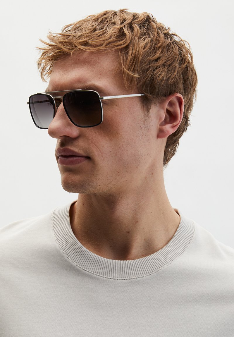 Jeune homme aux cheveux blonds courts et ébouriffés, portant des lunettes de soleil rectangulaires foncées et un t-shirt blanc à col rond, regardant vers sa gauche.