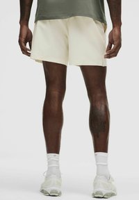 Mænds cremede shorts med en glat tekstur, parret med hvide sportsokker og lyse løbesko, set fra ryggen.