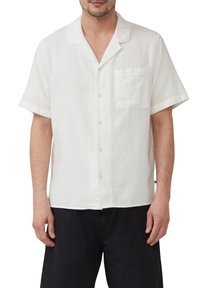 Witte kortegalm shirt met een klassieke kraag, gestructureerde stof, klein borstzakje en knoopsluiting aan de voorkant. G gedragen met een donkere broek.