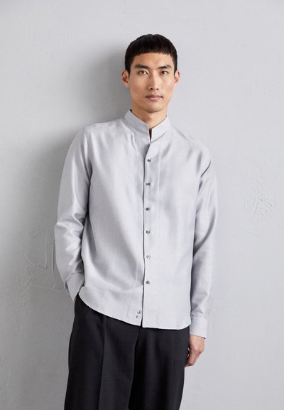 Camisa de patrón gris claro con cuello alzado, cierre de botones en la parte delantera, mangas largas y un corte holgado, combinada con pantalones oscuros.