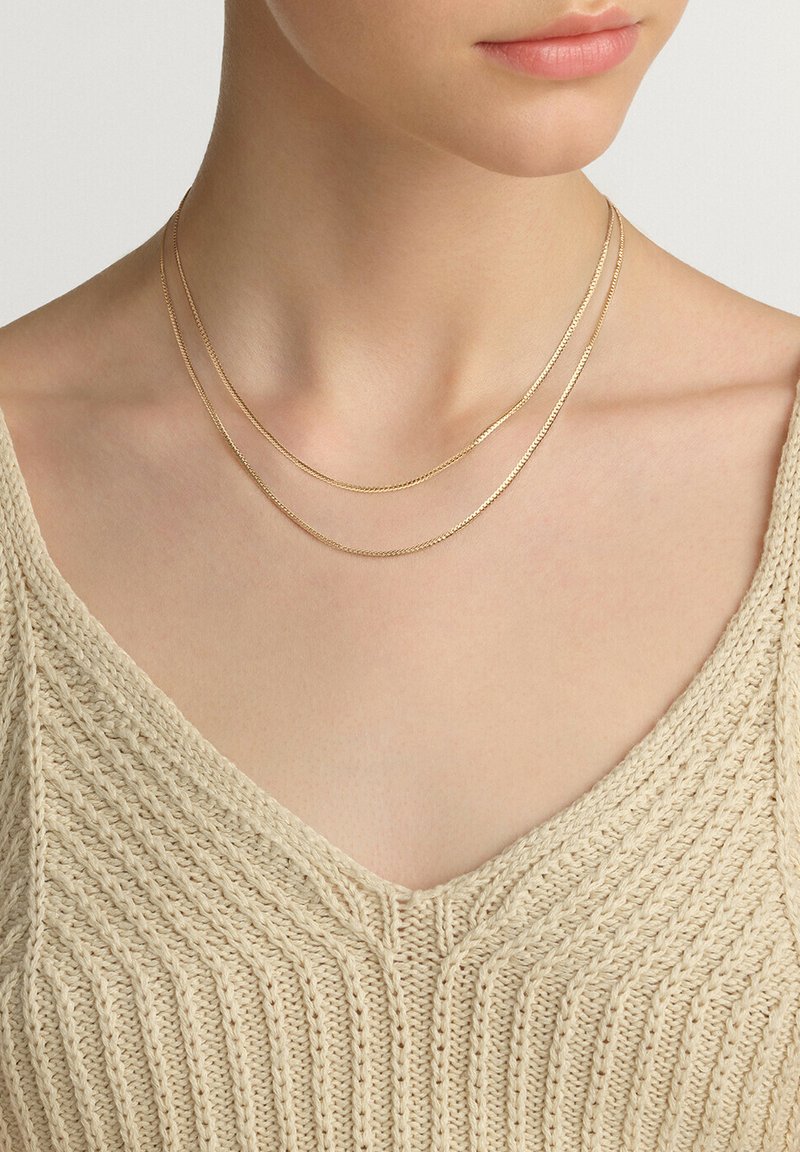 Collares en capas de oro presentan texturas variadas con eslabones lisos y retorcidos, descansando sobre una blusa de punto texturizada en color beige con escote en V.