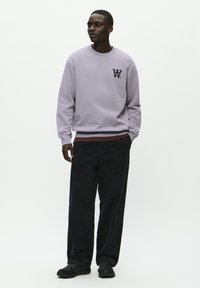 Light purple sweatshirt with crew neck and logo, paired with black corduroy pants and black sneakers, model standing against a white background.