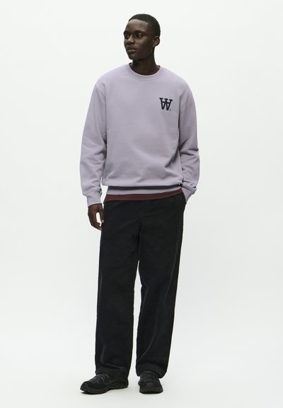 Sweat-shirt lilas clair avec col rond et logo, associé à un pantalon en velours côtelé noir et des baskets noires, mannequin se tenant contre un fond blanc.