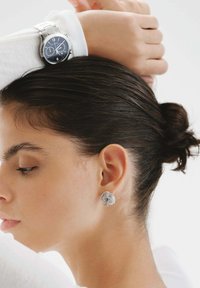 Se pueden ver pendientes en espiral de plata y un reloj de acero inoxidable con un dial oscuro. El cabello está recogido en un moño bajo frente a un fondo neutro.