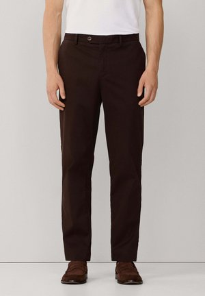 Hombre con pantalones marrón oscuro de pierna recta, camisa blanca metida por dentro y mocasines de gamuza marrón, de pie contra un fondo liso.