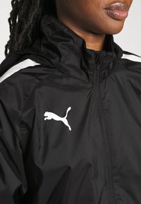 Sort vindjakke med høj krave, der har hvide sidelængder og et hvidt Puma-logo. Lavet af letvægtigt, struktureret stof.
