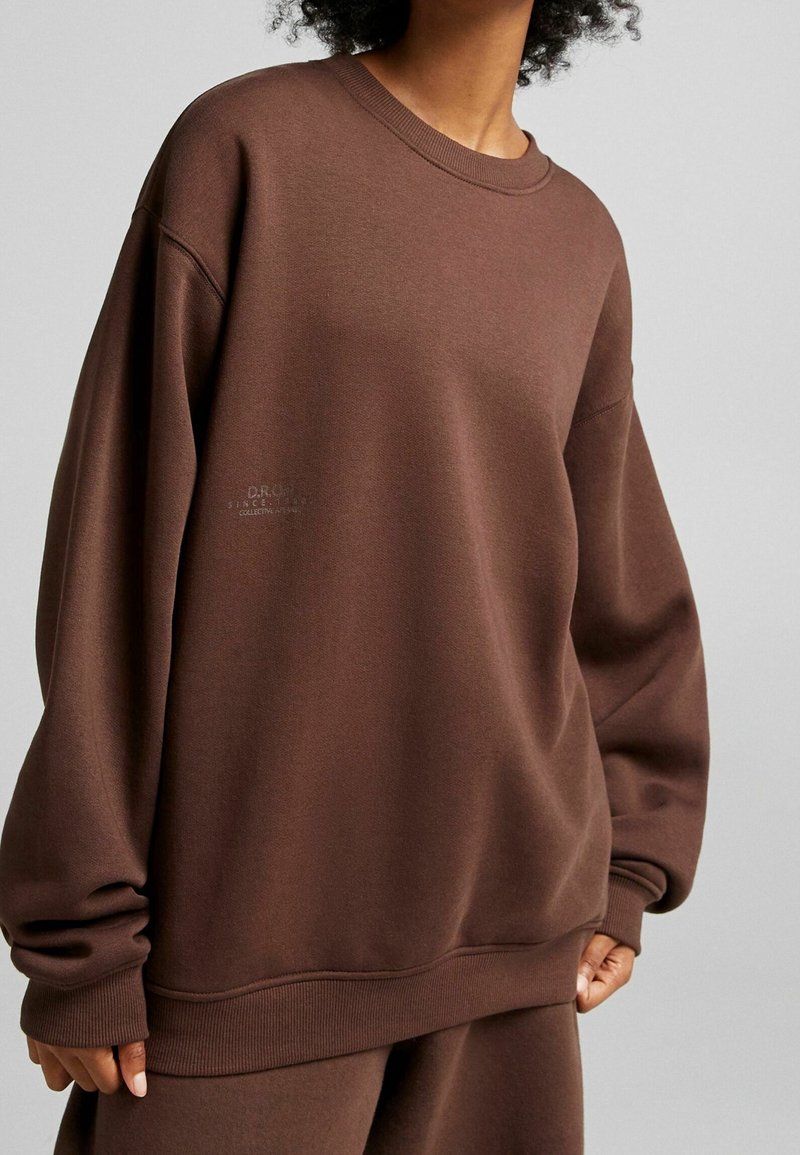 Brun oversized sweatshirt tillverkad av texturerad tyg med ribbad rund hals, breda ärmar och subtil märkning nära fållen.