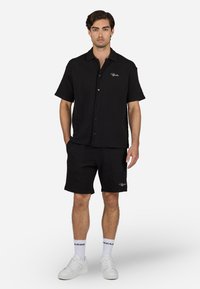 Black short-sleeve shirt with diamond pattern and logo, paired with matching black shorts. White sneakers and socks complete the outfit.