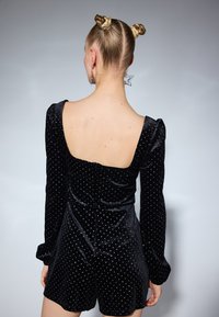 Mono de terciopelo negro con mangas largas, espalda cuadrada y estampado de topos. Presenta una textura suave y un diseño ajustado, que termina a medio muslo.