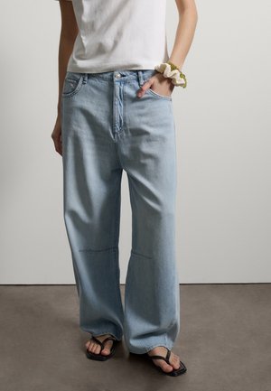 Persona che indossa jeans azzurri a gamba larga, t-shirt bianca, infradito nere e un elastico bianco e verde al polso.