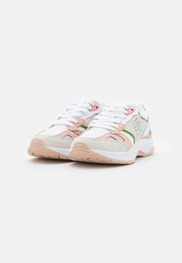 Des chaussures de sport blanches avec des accents roses et verts, présentant une surface texturée, un logo prominent et une semelle en caoutchouc flexible.