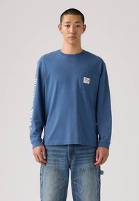 Jauns vīrietis ar zilu garām piedurknēm kreklu ar krūšu kabatu un baltu tekstu uz kreisās piedurknes, apvienojot ar zilajiem džinsiem.