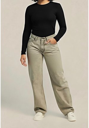 Kvinde iført sort langærmet top, beige bukser med høj talje og hvide sko, med den ene hånd i lommen, stående mod en ensfarvet baggrund.