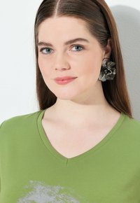 Camiseta verde de cuello en V con un acabado texturizado, que presenta un patrón abstracto gris. Grandes pendientes florales negros intrincados complementan el look.