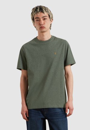 Giovane uomo con capelli biondi corti che indossa una t-shirt verde oliva a maniche corte e jeans blu su uno sfondo chiaro e uniforme.