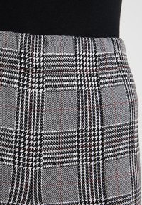 Tessuto a scacchi bianco e nero con motivi houndstooth e tartan, caratterizzato da accenti rossi. Texture morbida, design sartoriale e forma strutturata.