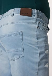 Vaqueros de mezclilla de color azul claro con una etiqueta de cuero marrón que dice "MEN+", con una textura desgastada y dos bolsillos en la parte trasera.