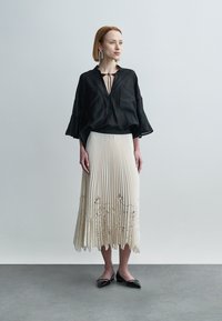 Μαύρη, άνετη μπλούζα με μανίκια καμπάνα, συνδυασμένη με πλισέ φούστα σε κρεμ χρώμα με κεντήματα λουλουδιών. Το μοντέλο φοράει μαύρα φλατ παπούτσια.